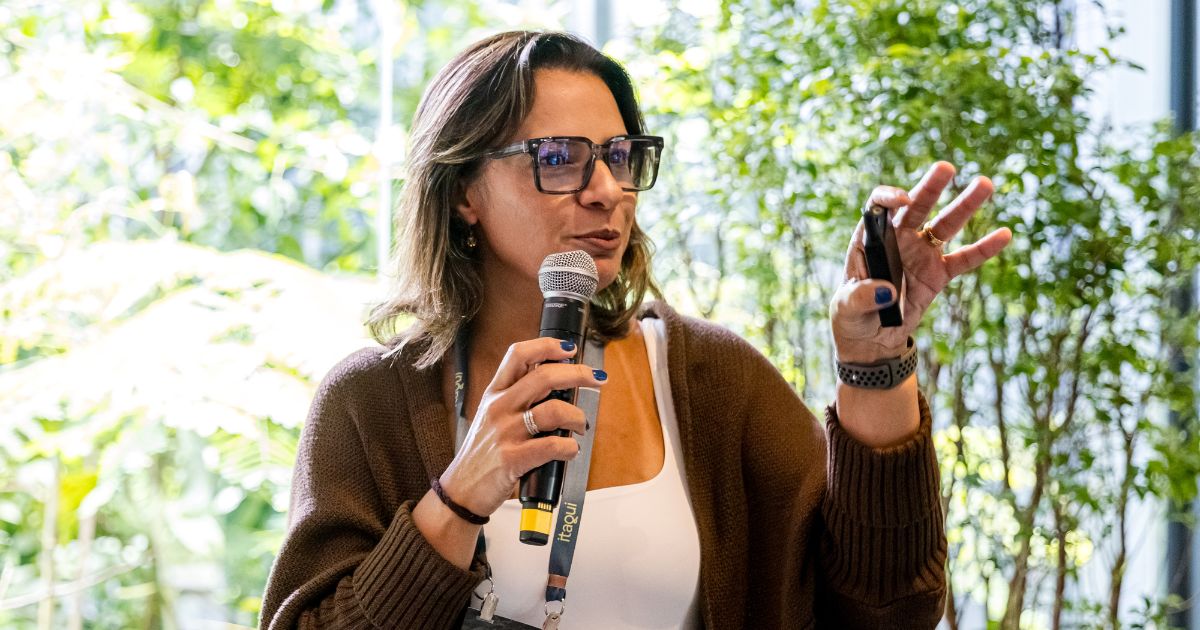 Uma pessoa está falando em um ambiente iluminado por luz natural, segurando um microfone em uma mão e um controle remoto de apresentação na outra. Ela veste um cardigã marrom sobre uma blusa clara e usa óculos. Ao fundo, há plantas verdes que compõem um cenário natural e suave. (mudança)