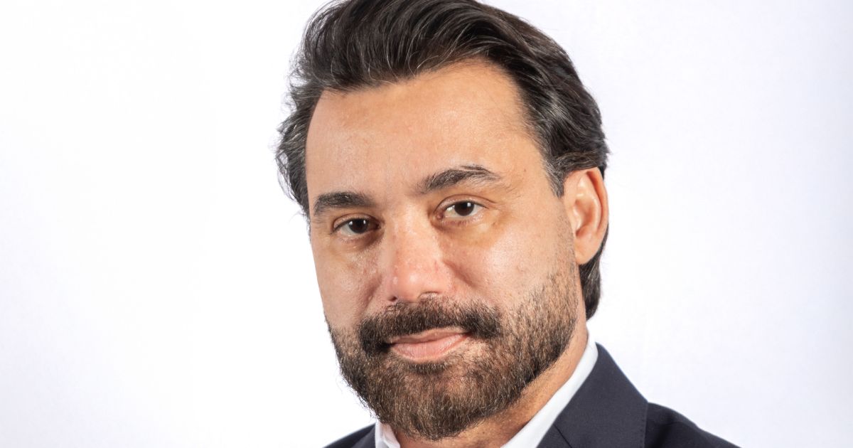 Uma pessoa vestindo um terno escuro com camisa branca aparece em um retrato profissional diante de um fundo totalmente branco. A imagem é enquadrada da altura do peito para cima, destacando o traje formal e o cabelo escuro cuidadosamente arrumado. (Approach Tech)