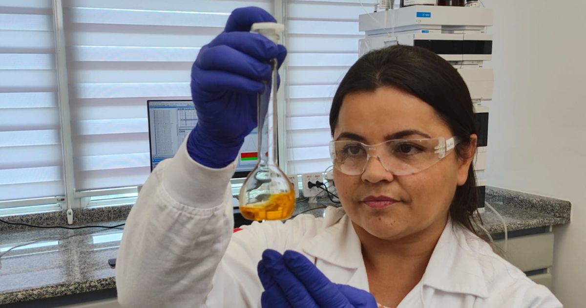 Uma pessoa usando jaleco branco e luvas azuis de laboratório segura um frasco volumétrico de vidro contendo uma substância líquida amarela. Ela está em um ambiente de laboratório, com bancadas, equipamentos analíticos e uma janela com persianas ao fundo. A ação sugere manipulação ou análise de amostras químicas. (mulheres)