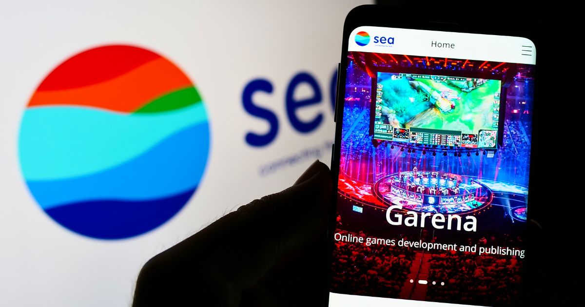 Um smartphone exibe a página da Garena, mostrando uma arena de e‑sports iluminada com telões transmitindo uma partida. Ao fundo, aparece ampliado o logotipo colorido da Sea, composto por faixas curvas em tons de azul, verde, vermelho e laranja. A composição destaca a relação entre a empresa Sea e sua divisão de jogos, Garena.