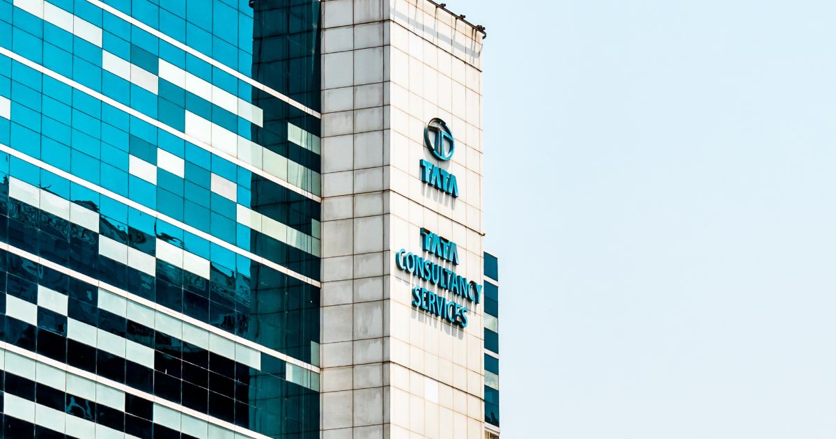 Um edifício corporativo alto, com fachada composta por painéis de vidro azulados e elementos geométricos, exibe o logotipo e o nome “Tata Consultancy Services” em letras azuis na torre central. O céu ao fundo está claro e sem nuvens, destacando a estrutura moderna da construção. (TCS)