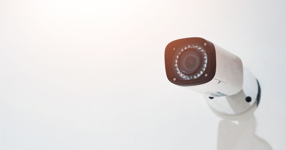 Uma câmera de segurança fixa está posicionada em uma parede clara, voltada para frente. O equipamento possui corpo branco e lente central cercada por LEDs infravermelhos, sugerindo capacidade de gravação mesmo em baixa luminosidade. A iluminação suave no canto superior esquerdo cria um clima minimalista e clean. (vigilância)