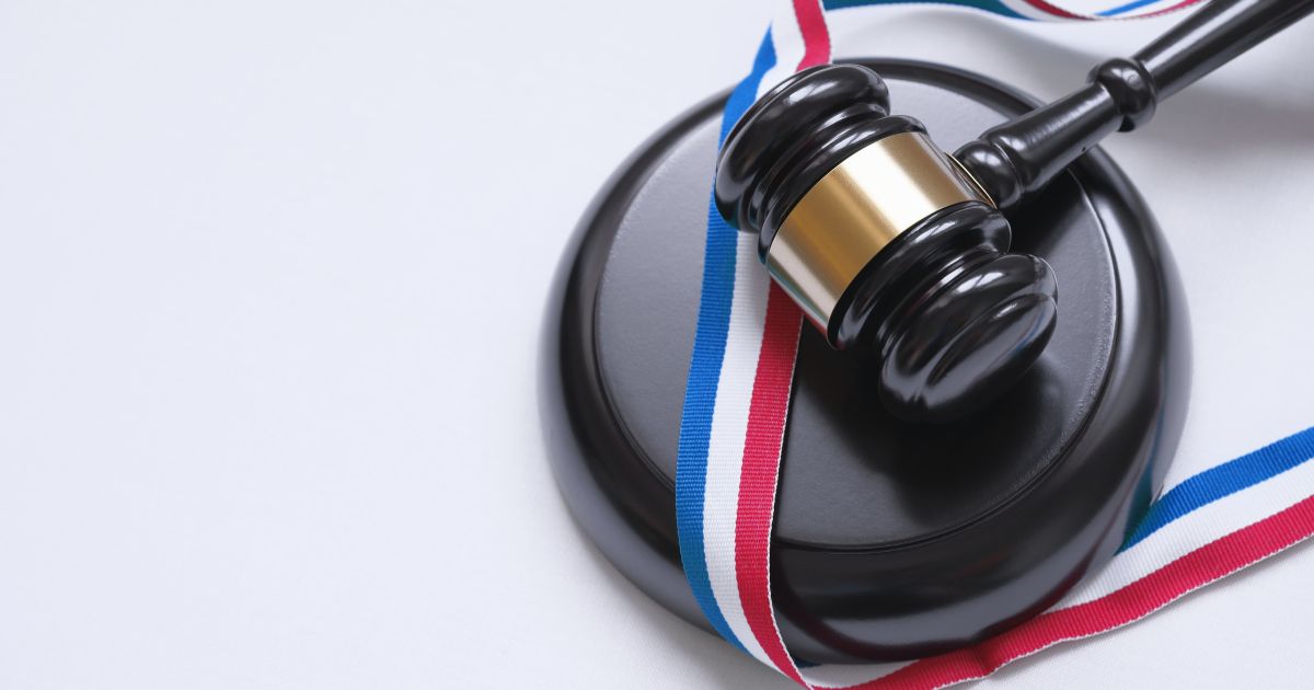 Um martelo de juiz repousa sobre uma base redonda escura, acompanhado por uma fita tricolor nas cores azul, branca e vermelha que se estende ao redor do conjunto. A cena remete ao universo jurídico e à tomada de decisões formais em um tribunal. (X)