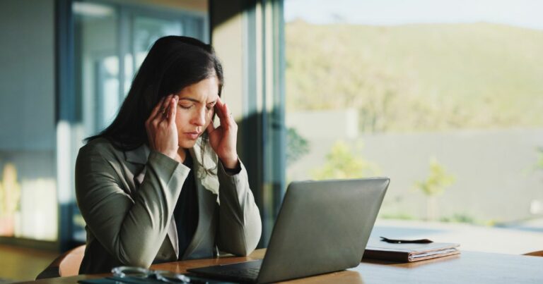 Uma pessoa sentada à mesa em um ambiente iluminado pela luz natural segura a cabeça com ambas as mãos, em postura que sugere preocupação ou cansaço. À sua frente, há um notebook aberto, um caderno e um par de óculos sobre a mesa. O fundo mostra janelas amplas com vista para uma área externa verde e ensolarada. (Saúde mental trabalho)