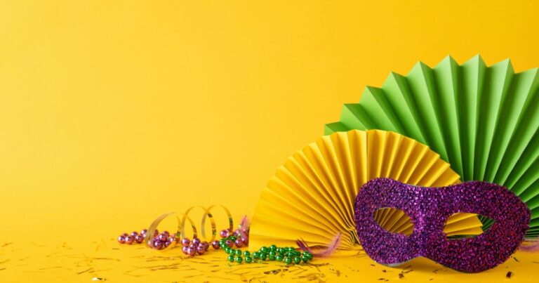 Itens de festa coloridos estão dispostos sobre um fundo amarelo vibrante. À direita, há uma máscara roxa com glitter, e atrás dela dois leques de papel em amarelo e verde. No canto esquerdo inferior, colares de contas metálicas em tons de roxo, dourado e verde aparecem espalhados, junto com serpentinas e pequenos confetes. A composição transmite clima de celebração, típico de festividades como Carnaval. (bloco)