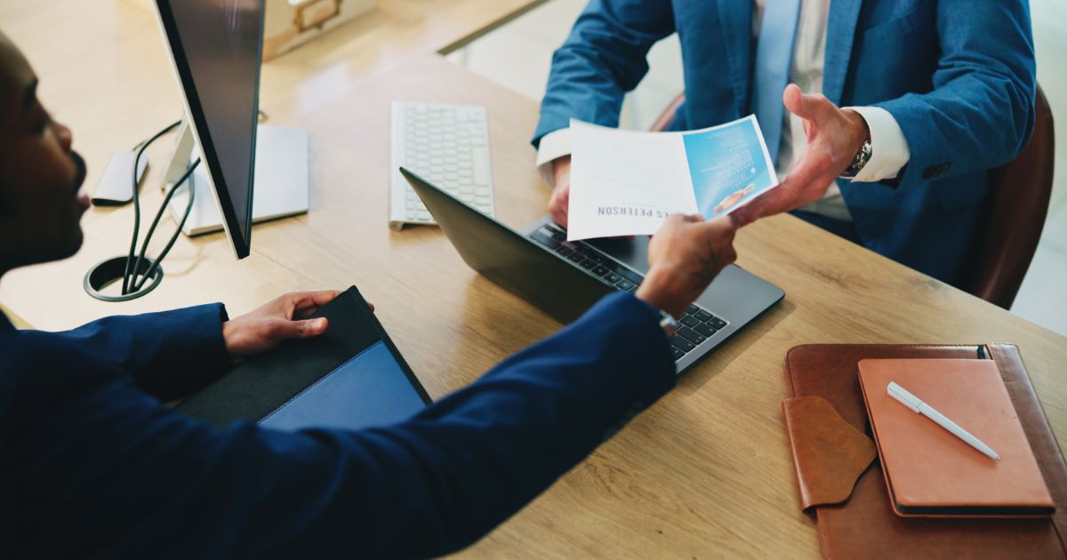 Duas pessoas em trajes formais interagem em uma mesa de reunião. Uma delas entrega um documento impresso com gráficos, enquanto a outra estende a mão para recebê‑lo. Sobre a mesa há um laptop aberto, um notebook de couro com caneta, um teclado externo e outros materiais de trabalho. O ambiente sugere uma negociação, revisão de contrato ou apresentação profissional. (mercado)