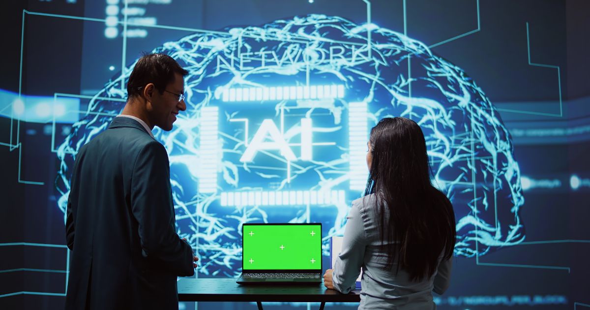 Duas pessoas observam uma grande projeção digital que exibe a ilustração de um cérebro formado por circuitos e linhas luminosas em azul, com a palavra “AI” ao centro. À frente delas, há um notebook aberto sobre uma mesa, com a tela em verde sólido, sugerindo uso de chroma key. O ambiente tem aparência tecnológica e imersiva, como uma demonstração ou apresentação de sistemas de inteligência artificial. (